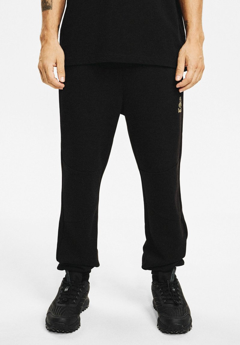 Mężczyzna w czarnych spodniach dresowych i czarnych trampkach, stojący z rękami swobodnie opartymi wzdłuż ciała na białym tle.