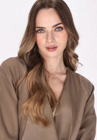 Brun satin blazer med sjalkrave og flydende stof, båret over en lys top. Lange, bølgende hår og en neutral makeup palette.