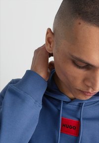 Hoodie in cotone blu con una pattina rettangolare rossa sul davanti che mostra la scritta "HUGO". Presenta un cappuccio con coulisse e polsini a coste. Vista ravvicinata.