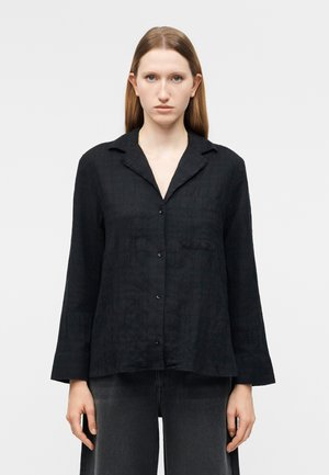 Kobieta o prostych brązowych włosach, ubrana w luźną czarną koszulę na guziki i ciemnoszare spodnie, stojąca na tle jednolitego jasnego tła.