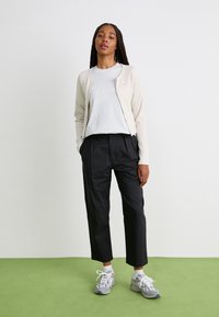 Cardigan côtelé beige clair sur un t-shirt gris, associé à un pantalon noir court et des baskets grises. Sur un fond uni.