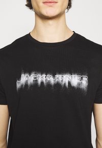 Svart bomull t-shirt med en blekt vit grafisk tryck av texten "JACK&JONES" över bröstet och rund halsringning.
