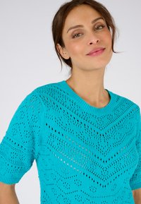 Suéter de punto de color turquesa con mangas cortas, que presenta un patrón de formas de diamantes y chevrón, y un cuello acanalado.