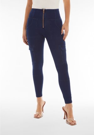 Nainen, joka käyttää tummansiniset korkeavyötäröiset kapeat housut, joissa on etuvetoketju ja sivutaskut, yhdistettyinä läpinäkyvillä korollisilla sandaaleilla.