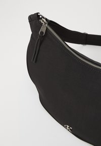 Czarna nylonowa torba na ramię z metalicznym srebrnym zamkiem błyskawicznym i logo, o gładkiej fakturze oraz zaokrąglonym, kompaktowym kształcie.