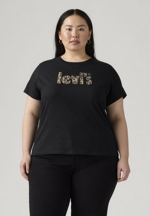 Vrouw met lang donker haar die gouden oorringen draagt en een zwart Levi's T-shirt met een luipaardprint-logo op een effen grijze achtergrond.