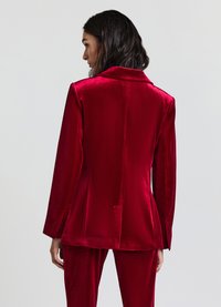 Blazer en velours rouge avec une coupe structurée, un col cranté, des manches longues et une texture lisse. Il présente une fente unique au dos.