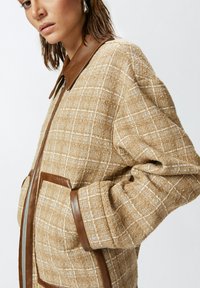 Chaqueta marrón con textura y cremallera frontal que presenta un patrón de cuadros, cuello de cuero marrón y ribete, y dos bolsillos delanteros.