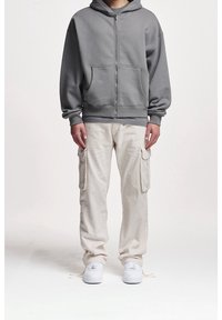 Šedá zip-up mikina s velkými předními kapsami, v kombinaci s béžovými cargo kalhotami s bočními kapsami. Bílé boty dotvářejí outfit.