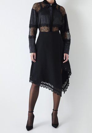 Musta mekko, jossa on puoliläpinäkyvä pitsiyläosa kauluksella ja pitkät hihat, yhdistettynä istuvaan hameeseen, joka päättyy kaarevaan helmaukseen.
