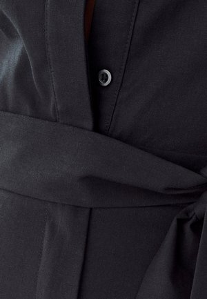 Melnas krekla detaļas, kurās redzama auduma tekstūra, pogu aizdare un auduma josta. Dizains ietver gludu izskatu un slāņotas auduma daļas.