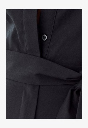 Detail černé košile se zaměřením na texturu látky, zapínání na knoflíky a látkový pásek. Design zahrnuje hladký vzhled a vrstvené části látky.