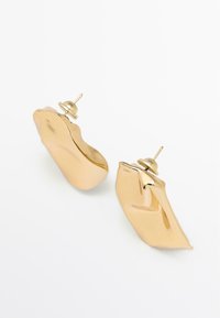 Boucles d'oreilles en métal doré avec un design ondulé et abstrait. Fermeture sécurisée avec une petite tige. Surface lisse et réfléchissante.