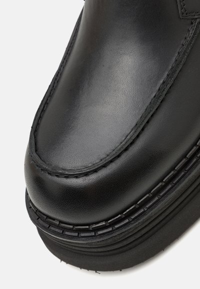 Mocassins en cuir noir avec une surface lisse, un bout arrondi, des coutures visibles et une semelle en caoutchouc noir épaisse avec des accents texturés.