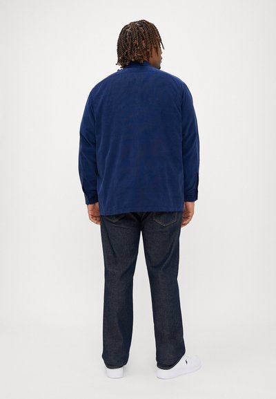 Ναυτικό μπλε πουκάμισο από χοντρή βελόνα με γιακά, μακριά μανίκια και κουμπιά στους καρπούς, φορεμένο με σκούρο τζιν παντελόνι και λευκά αθλητικά παπούτσια.