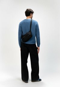 Borsa a tracolla nera con parte superiore arricciata, indossata sopra un maglione blu e pantaloni a gamba larga neri. Texture liscia, design minimalista e tracolla regolabile.