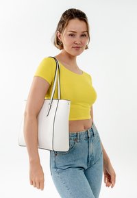 Sac fourre-tout blanc avec des bordures noires, fabriqué en matériau texturé. Modèle portant un haut court jaune et un jean taille haute bleu. Design simple.