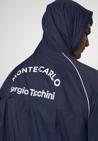 Tumši zils kapuces jaka ar baltu tekstu "MONTE-CARLO" un "Sergio Tacchini" aizmugurē, ar gludu faktūru un brīvu piegriezumu.