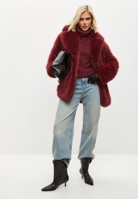 Abrigo de piel sintética burdeos con mangas anchas, suéter de punto burdeos de cuello alto, jeans holgados de un azul claro y botas negras con puntera y tacones.