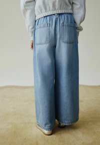 Plave traperice širokih nogavica s visokim strukom, elastičnom trakom i dva stražnja džepa, prikazane s leđa na bež podlozi.