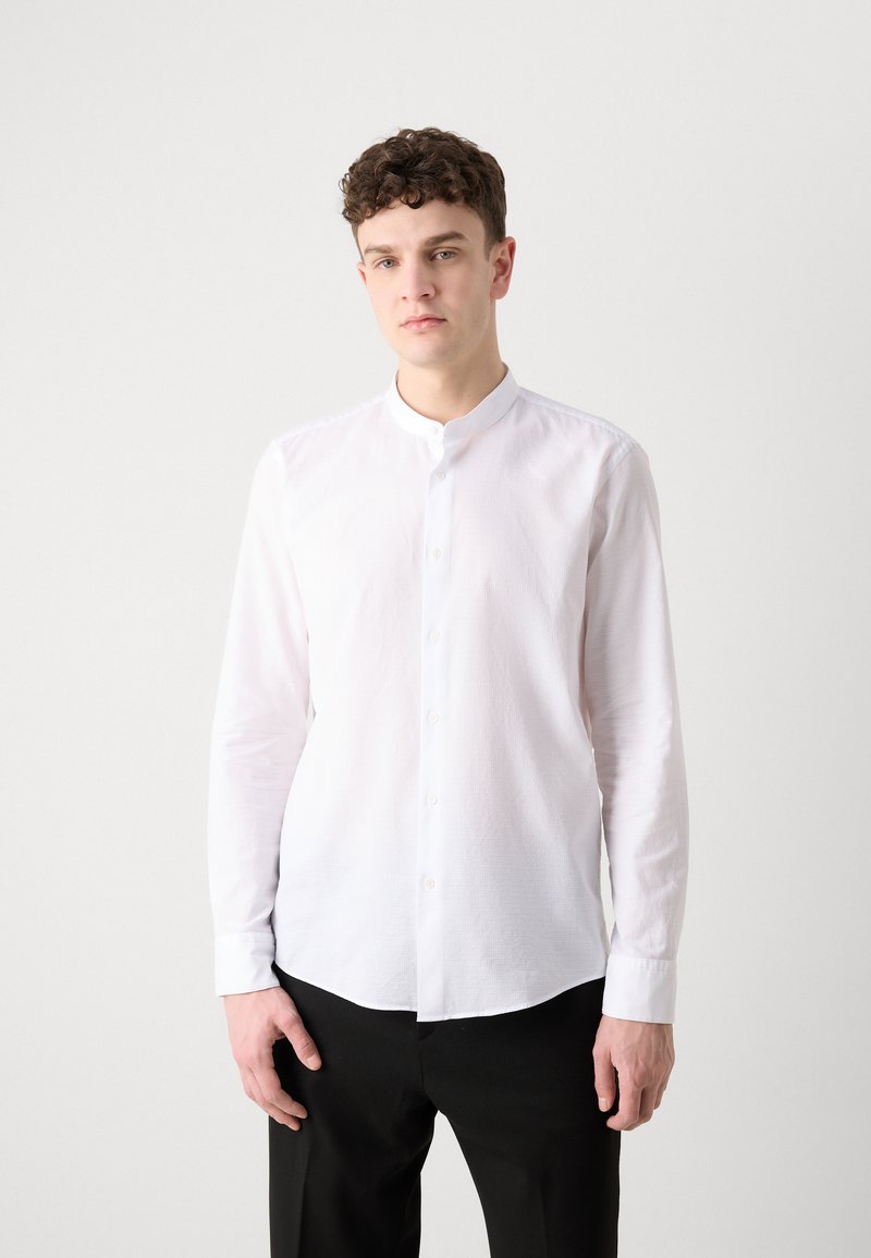 Biała koszula z długim rękawem i kołnierzykiem mandarynki, wykonana z lekkiego materiału. Posiada zapięcie na guziki z przodu oraz subtelnie teksturowaną powierzchnię. Czarne spodnie.