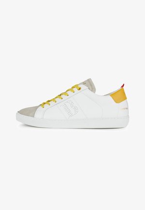 Baskets basses blanches avec lacets jaunes, bout en daim beige, languette de talon jaune et logo perforé sur le côté.