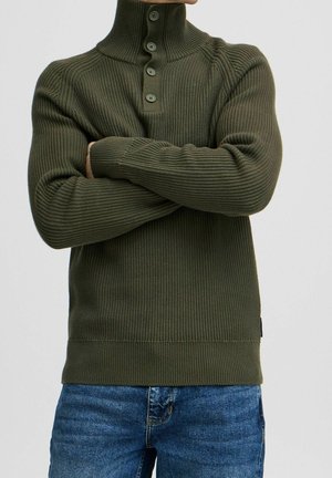 Personne portant un pull en maille côtelée vert olive avec un col montant et quatre boutons, les bras croisés sur la poitrine, associé à un jean bleu.