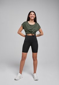 Olive green cropped top, knotted at the front, paired with high-waisted black bike shorts, and white athletic shoes. Smooth fabric, relaxed fit.