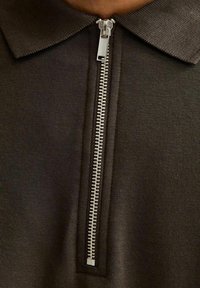 Polo di colore marrone scuro con chiusura a mezza zip dotata di una cerniera metallica, texture a maglia e design del colletto classico.