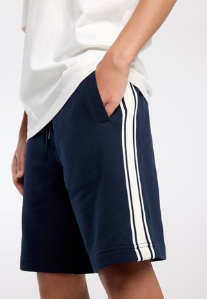 Persoon draagt marineblauwe korte broek met witte zijstrepen, hand in zak, gecombineerd met een losse witte blouse tegen een effen achtergrond.