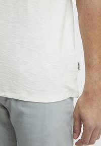 Kortærmet, lysegrå T-shirt med en tekstureret, let meleret indfarvning og et lille stofmærke ved sidesømmen. Sammen med grå bukser.