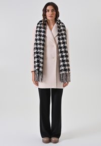 Dubbelgeknoopte lichtroze jas van gestructureerde stof, zwart-wit geruite sjaal, zwarte wijde broek en bruine instapschoenen.