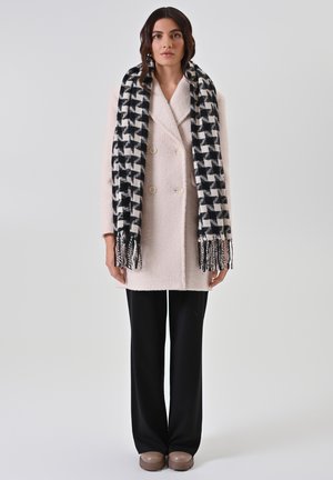 Dubbelgeknoopte lichtroze jas van gestructureerde stof, zwart-wit geruite sjaal, zwarte wijde broek en bruine instapschoenen.