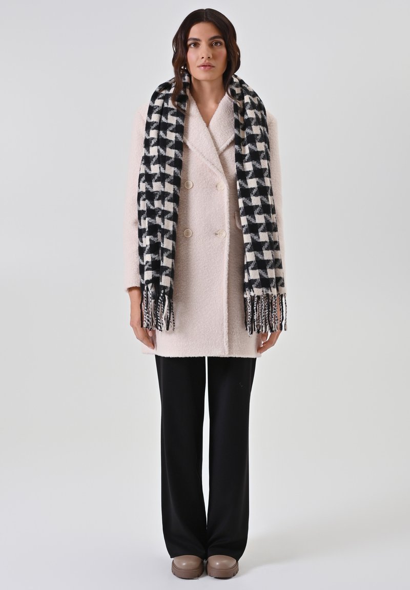 Dubbelgeknoopte lichtroze jas van gestructureerde stof, zwart-wit geruite sjaal, zwarte wijde broek en bruine instapschoenen.