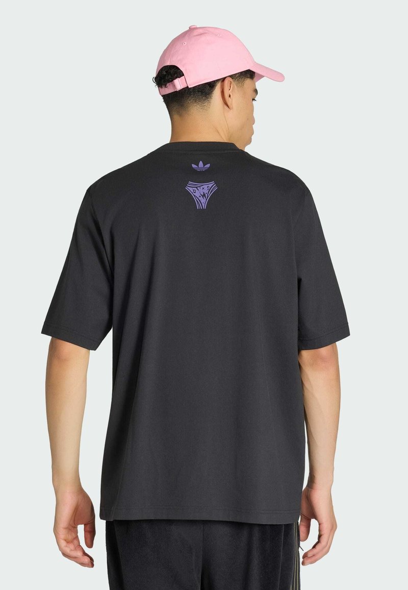 Mann trägt ein schwarzes übergroßes T-Shirt mit kleinem lila Adidas-Logo und einer Grafik auf dem oberen Rücken sowie eine rosa Baseballkappe, er ist von hinten zu sehen.