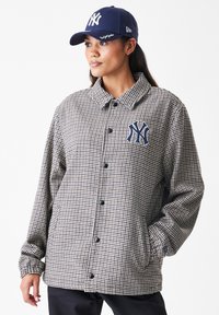 Γυναίκα που φοράει καπέλο ναυτικού New York Yankees και σακάκι με καρό σχέδιο, με το λογότυπο των Yankees στο στήθος, στέκεται με τα χέρια στις τσέπες.