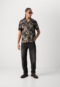 Camicia nera a maniche corte con un motivo astratto verde e grigio, abbinata a jeans scuri e stivali marroni. Tessuto morbido e vestibilità sartoriale.