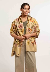 Châle fleuri en beige avec des motifs vibrants orange, rose et vert ; texture douce avec des franges sur les bords ; porté sur un haut vert et un pantalon beige.