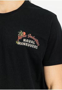 Černé bavlněné tričko s předním potiskem, na kterém je stylizovaný text "Alpha Industries" nad nápisem "NAVAL MANEUVERS", doplněné akcenty palmových stromů.