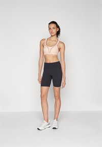 Lys fersken sports-bh med kontrasterende sømmer, sammen med høytlivede svarte sykkelshorts. Modellen har på seg hvite treningssko.
