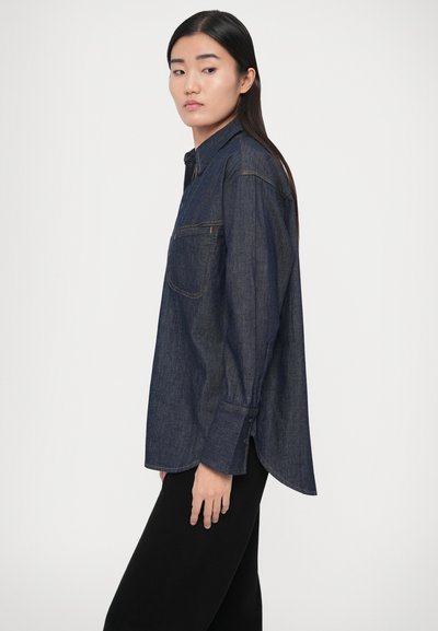 Džínová košeľa tmavomodrej farby s golierom, dlhými rukávmi a náprsným vreckom. Jemná textúra s viditeľnými stehmi.