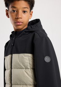 Black hooded jacket with a light olive green insulated panel. Features a round logo patch on the right sleeve. Smooth and textured materials.