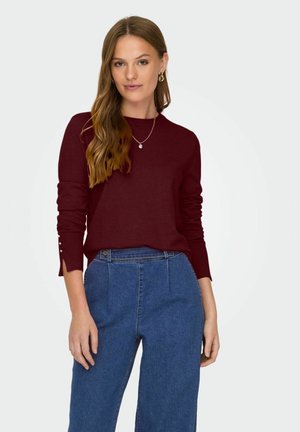Pull en maille bordeaux à manches longues, avec des accents de boutons aux poignets. Associé à un pantalon en denim bleu taille haute.