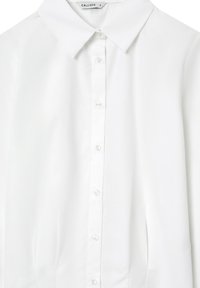 Camicia bianca con colletto realizzata in tessuto liscio, caratterizzata da una chiusura a bottoni e leggere pieghe in vita per dare forma. Bottoni trasparenti visibili.