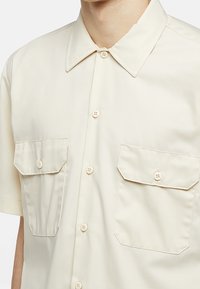 Camicia a maniche corte di colore beige chiaro, con colletto, due tasche frontali e una texture liscia e opaca.