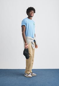 T-shirt azul claro com mangas dobradas, camisola cinzenta amarrada na cintura, calças bege e boné preto. Calçado com design cinzento e branco.