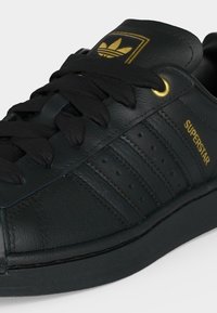 Μαύρο αθλητικό παπούτσι Adidas Superstar με χρυσό λογότυπο στη γλώσσα, χρυσή θηλιά και "SUPERSTAR" κείμενο σε χρυσό χρώμα στο πλάι κοντά στις τρεις υφασμάτινες ρίγες.