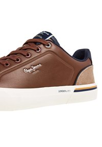 Zapatilla de deporte de cuero sintético marrón con detalles en ante color canela, suela de goma blanca, cordones marrones y marca en el lateral y la lengüeta.