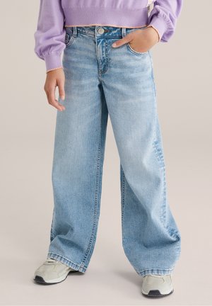 Personne portant un jean large bleu clair, des baskets blanches et un pull lavande avec une main dans la poche.