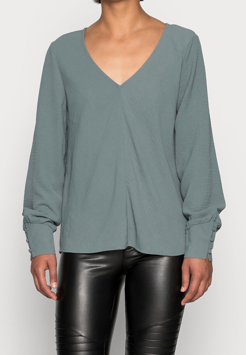 Kobieta w luźnej, teksturowanej, zakurzonej zielonej bluzce z dekoltem w serek, z guzikami na mankietach, oraz w błyszczących czarnych skórzanych spodniach, stojąca na białym tle.